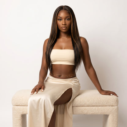 A woman with long, straight dark brown hair sits on a textured cream-colored bench against a plain off-white background. She is wearing a matching beige two-piece set consisting of a strapless tube top and a high-slit maxi skirt, accessorized with a thin gold chain waist belt.