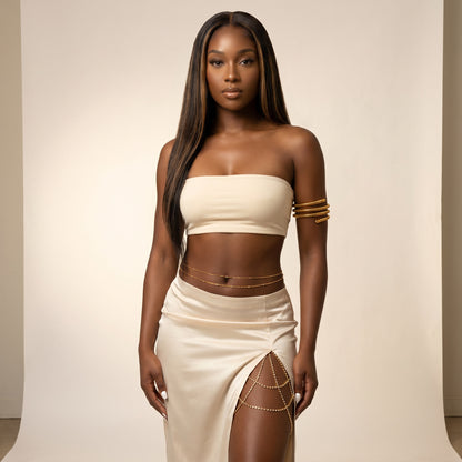 A woman modeling a cream-colored two-piece set consisting of a strapless bandeau crop top and a matching high-slit skirt, accessorized with gold chain details and bracelets, set against a plain beige studio backdrop.