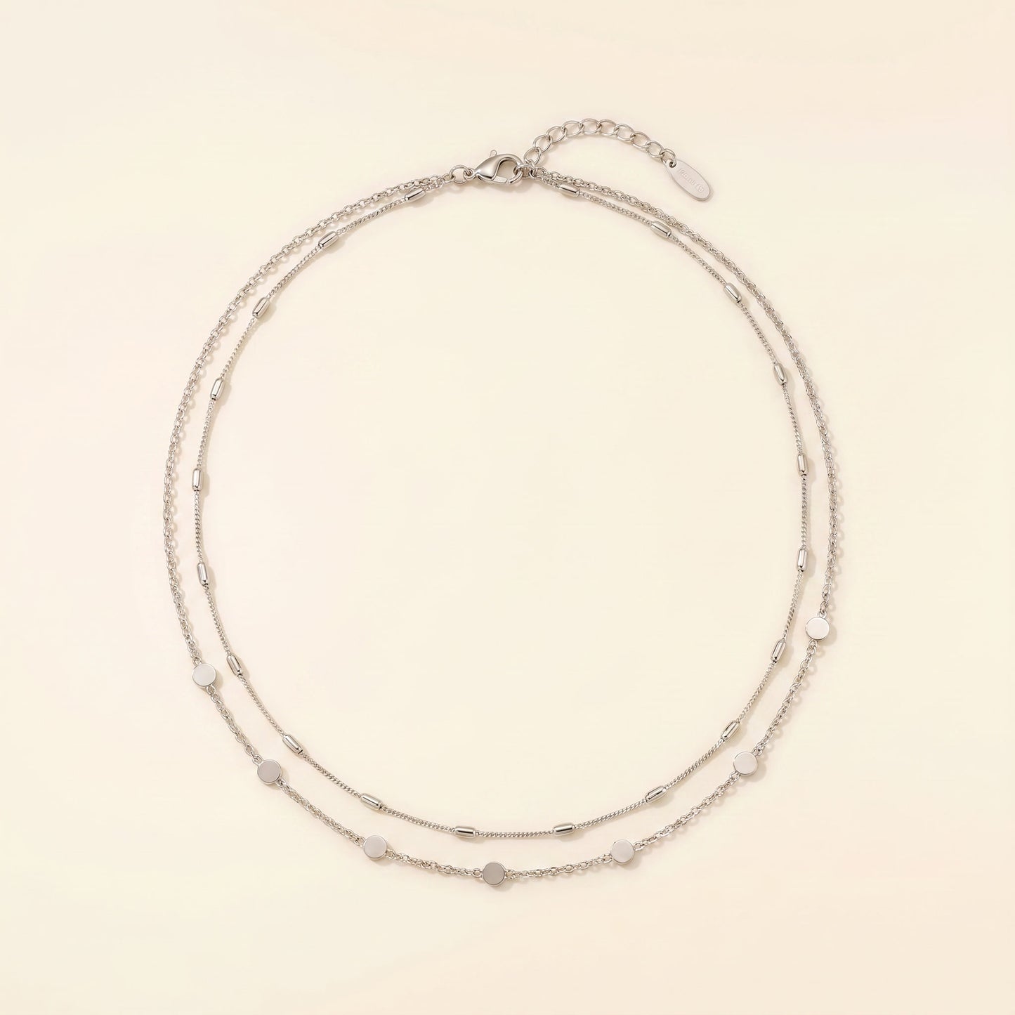 A delicate double-strand silver chain necklace with small, evenly spaced heart-shaped and oval beads, displayed on a warm cream background.