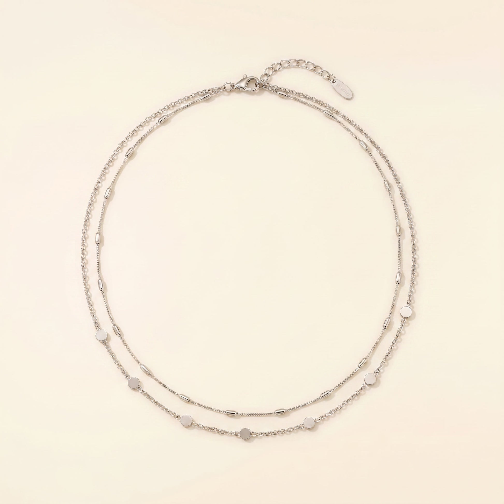 A delicate double-strand silver chain necklace with small, evenly spaced heart-shaped and oval beads, displayed on a warm cream background.