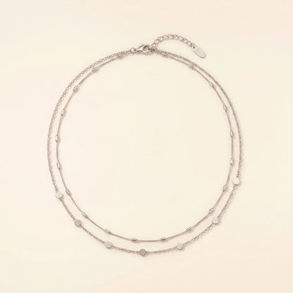 A delicate double-strand silver chain necklace with small, evenly spaced heart-shaped and oval beads, displayed on a warm cream background.