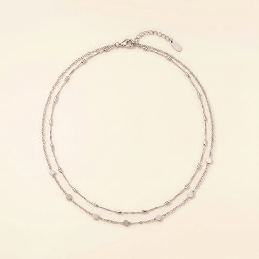A delicate double-strand silver chain necklace with small, evenly spaced heart-shaped and oval beads, displayed on a warm cream background.