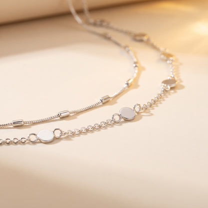 A delicate silver chain necklace with flat circular disc charms, displayed on a warm beige surface with a soft, warm-toned shadow.