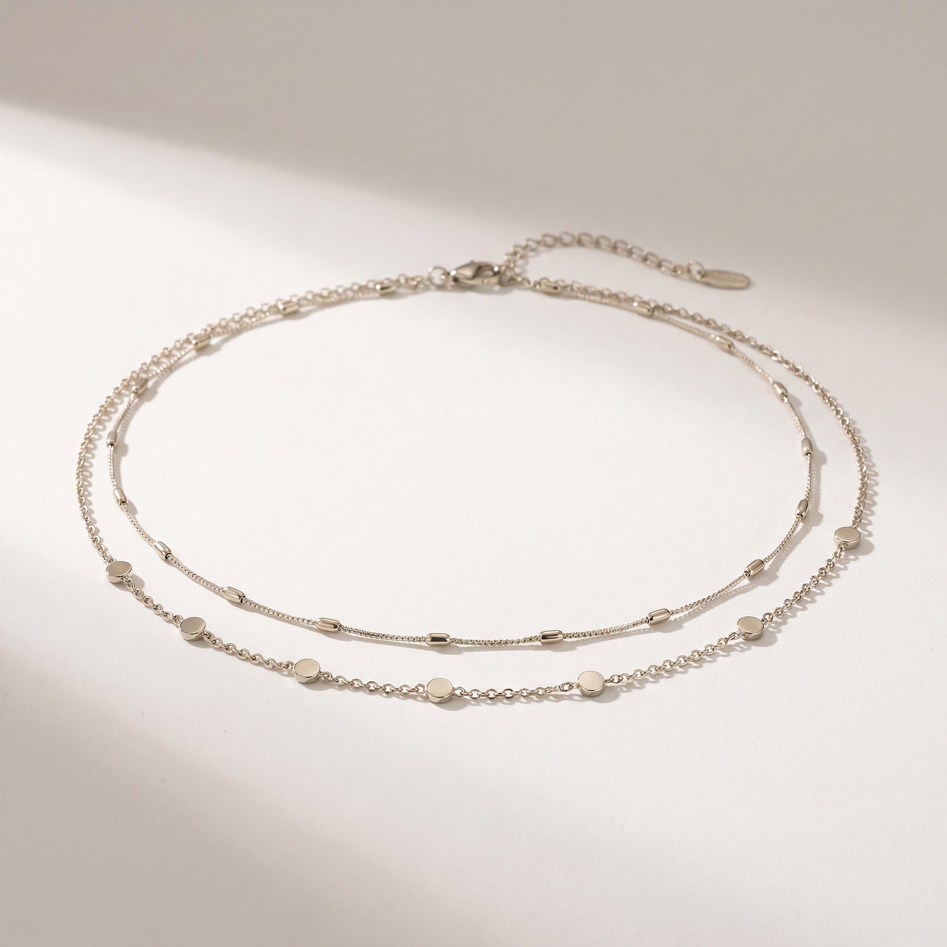 A delicate double-layered silver chain anklet with small flat disc charms, displayed on a clean white surface with soft shadows.