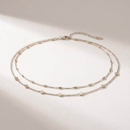 A delicate double-layered silver chain anklet with small flat disc charms, displayed on a clean white surface with soft shadows.
