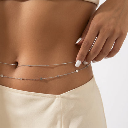 A close-up of a model's midsection wearing a delicate silver double-layered belly chain with small disc charms, paired with cream-colored bottoms.