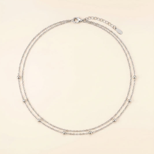 A delicate silver double-strand chain necklace with small metallic beads spaced along the chain, displayed on a light cream background.