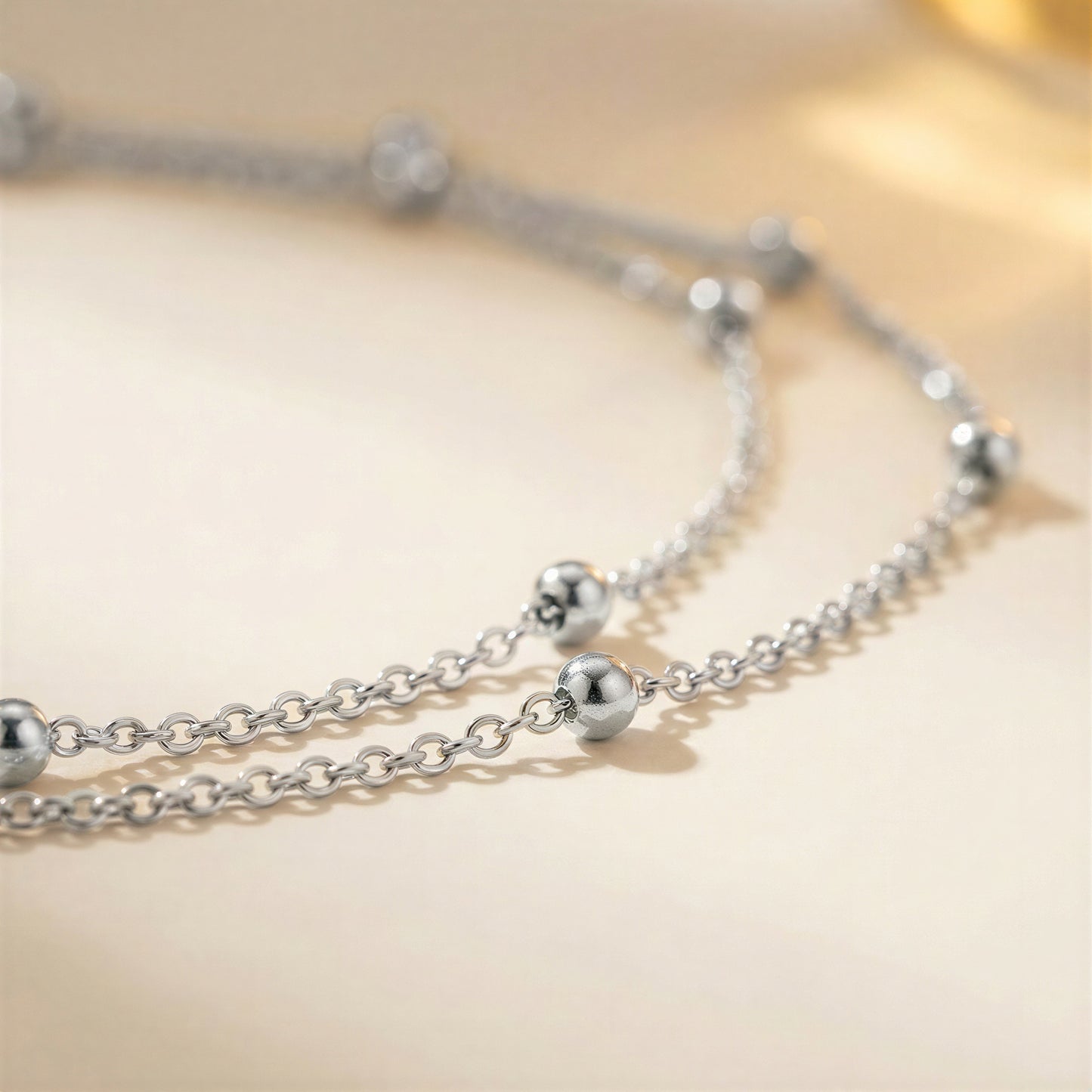 A close-up of a silver-toned double-strand chain necklace with polished spherical bead accents, resting on a soft beige surface.