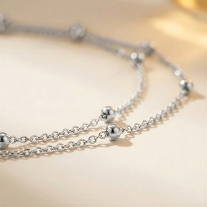 A close-up of a silver-toned double-strand chain necklace with polished spherical bead accents, resting on a soft beige surface.