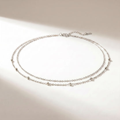 A delicate double-strand silver chain necklace with small bead accents, displayed on a clean white surface with soft shadows.