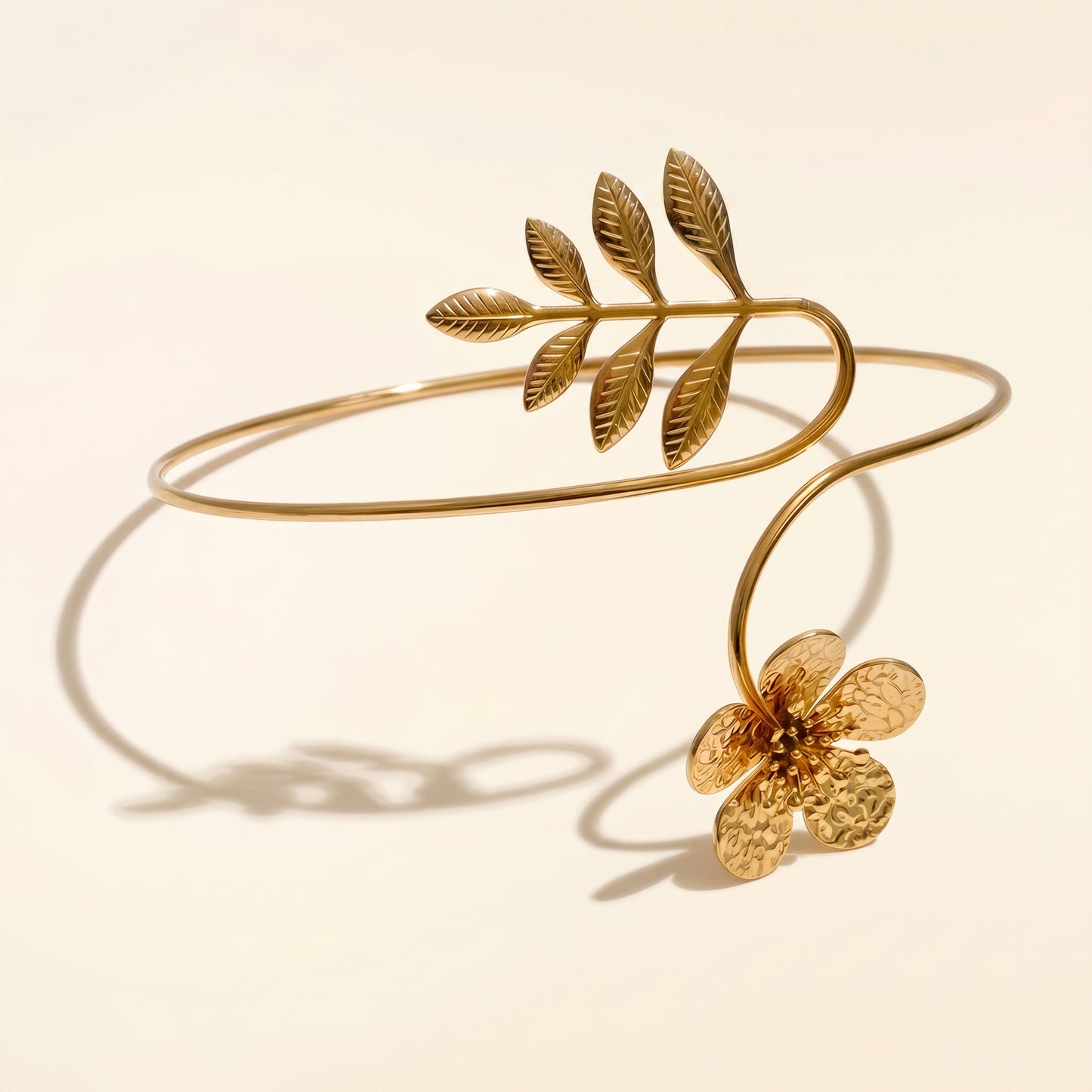 Gold-toned open bangle bracelet featuring a leafy branch motif and a textured flower detail, photographed on a warm cream background with a soft shadow.