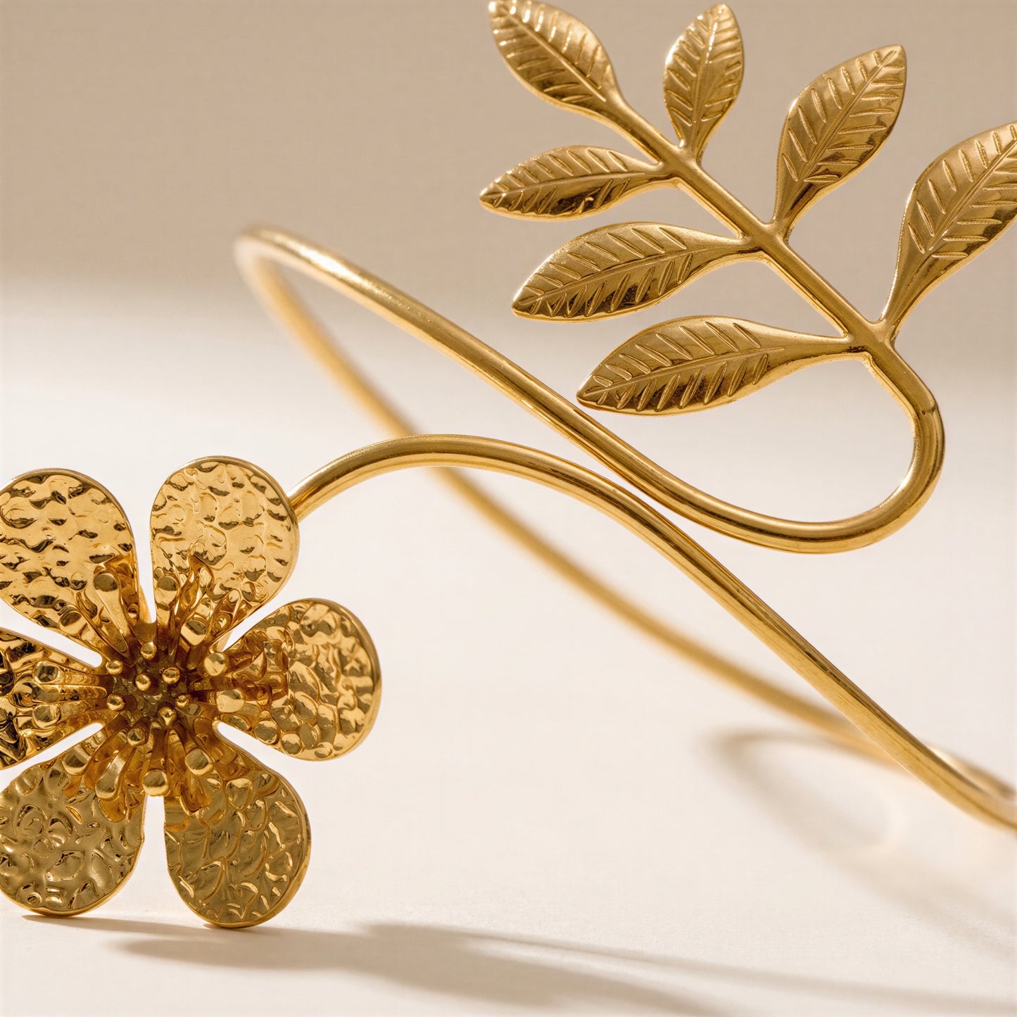 Gold-tone metal cuff bracelet featuring a textured flower on one end and a leafy branch on the other, photographed on a clean light beige background.