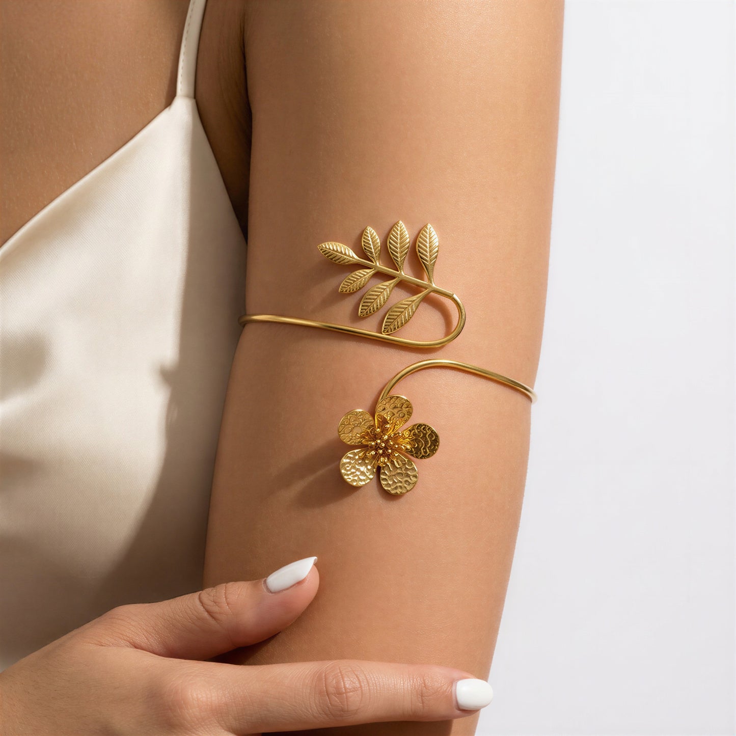 A gold leaf and flower cuff bracelet worn on a model's upper arm, shown against a light gray background with a cream satin top.