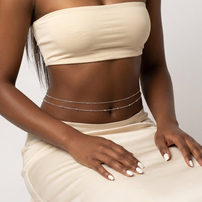 A woman modeling a cream-colored two-piece set consisting of a strapless bandeau crop top and a matching high-slit skirt, accessorized with gold chain details and bracelets, set against a plain beige studio backdrop.