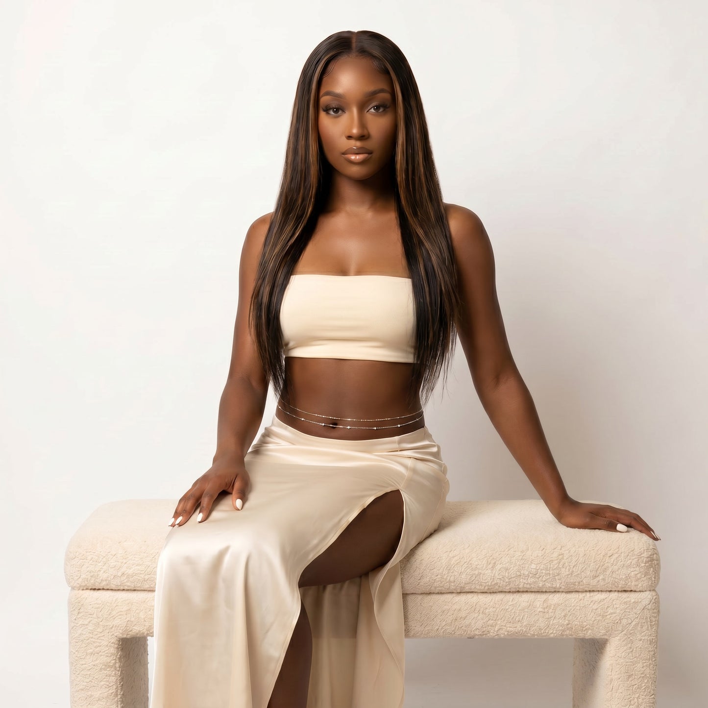 A woman with long, straight dark brown hair sits on a textured cream-colored bench against a plain off-white background. She is wearing a matching beige two-piece set consisting of a strapless tube top and a high-slit maxi skirt, accessorized with a thin gold chain waist belt.