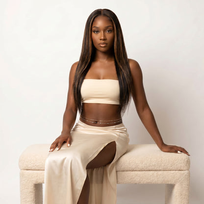 A woman with long, straight dark brown hair sits on a textured cream-colored bench against a plain off-white background. She is wearing a matching beige two-piece set consisting of a strapless tube top and a high-slit maxi skirt, accessorized with a thin gold chain waist belt.