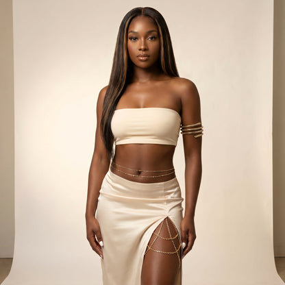 A woman modeling a cream-colored two-piece set consisting of a strapless bandeau crop top and a matching high-slit skirt, accessorized with gold chain details and bracelets, set against a plain beige studio backdrop.