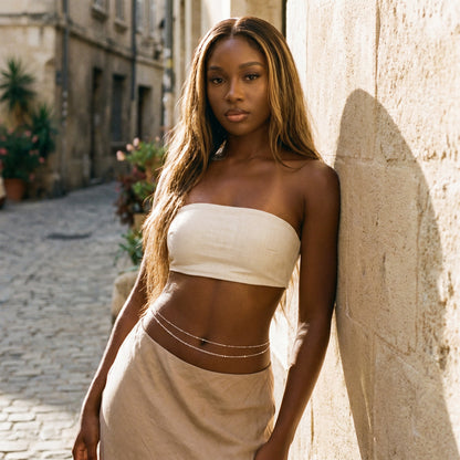 A woman with long light brown hair stands outdoors against a sunlit stone wall, wearing a cream strapless bandeau-style top and a beige skirt or drape, with thin chain accessories at the waist.