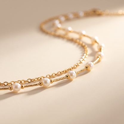 A delicate gold chain necklace with evenly spaced white pearl beads, photographed on a warm beige background with soft shadows.
