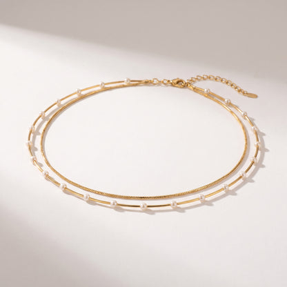 A delicate double-layered gold chain necklace with small white pearl accents, displayed on a clean, light beige background with soft shadows.