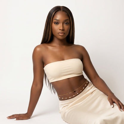 A woman posing in a studio setting wearing a cream-colored strapless tube top and a matching cream skirt with a thin brown waist chain.