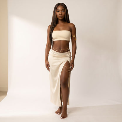 A woman stands barefoot in a studio wearing a cream two-piece outfit consisting of a strapless bandeau top and a matching high-slit maxi skirt, styled with gold chain and stud accessories.