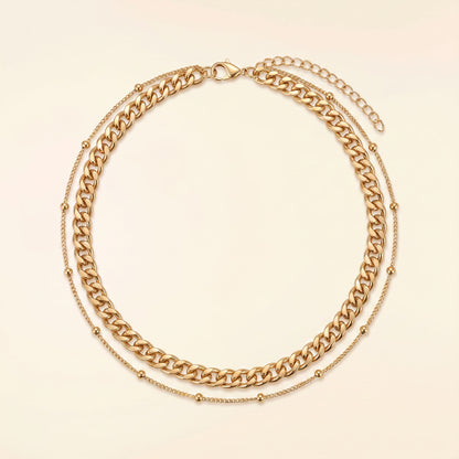 A gold-toned double-layer necklace featuring a chunky curb chain and a delicate beaded chain, displayed on a light cream background.