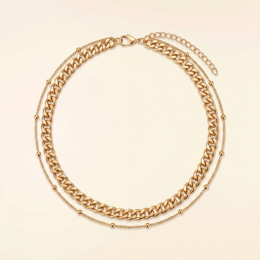 A gold-toned double-layer necklace featuring a chunky curb chain and a delicate beaded chain, displayed on a light cream background.