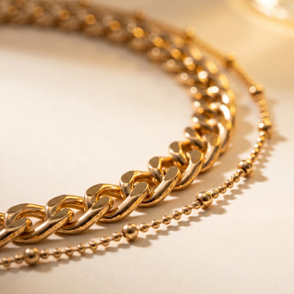 A gold-toned double-layer necklace featuring a chunky curb chain and a delicate beaded chain, displayed on a light cream background.