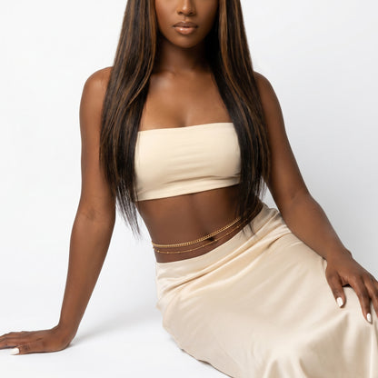 A woman wearing a beige strapless tube top and matching beige satin skirt, accessorized with a delicate gold chain belt, posed against a clean white studio background.