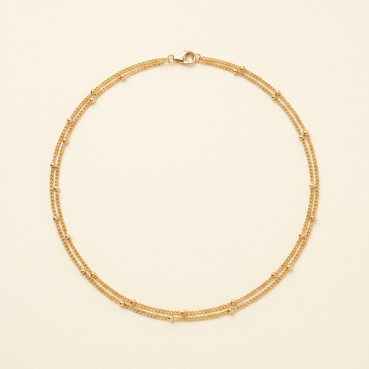 A delicate gold double-strand chain necklace with small bead accents, displayed on a light cream background.
