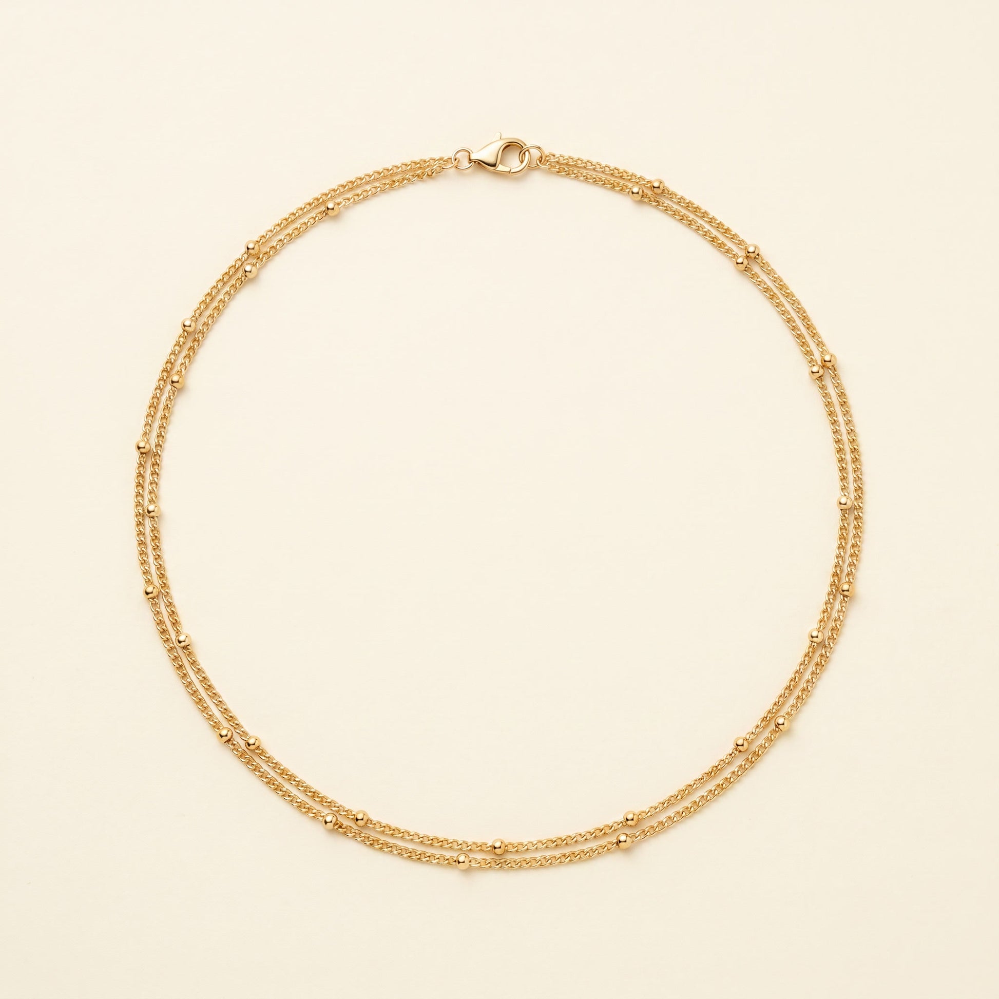 A delicate gold double-strand chain necklace with small bead accents, displayed on a light cream background.