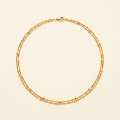 A delicate gold double-strand chain necklace with small bead accents, displayed on a light cream background.