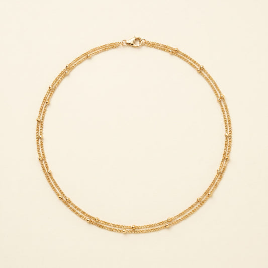 A delicate gold double-strand chain necklace with small bead accents, displayed on a light cream background.