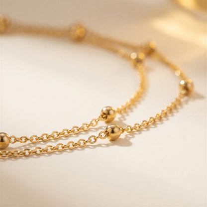 A close-up of a delicate gold chain necklace featuring small polished gold beads spaced along the chain, resting on a soft, light beige surface.