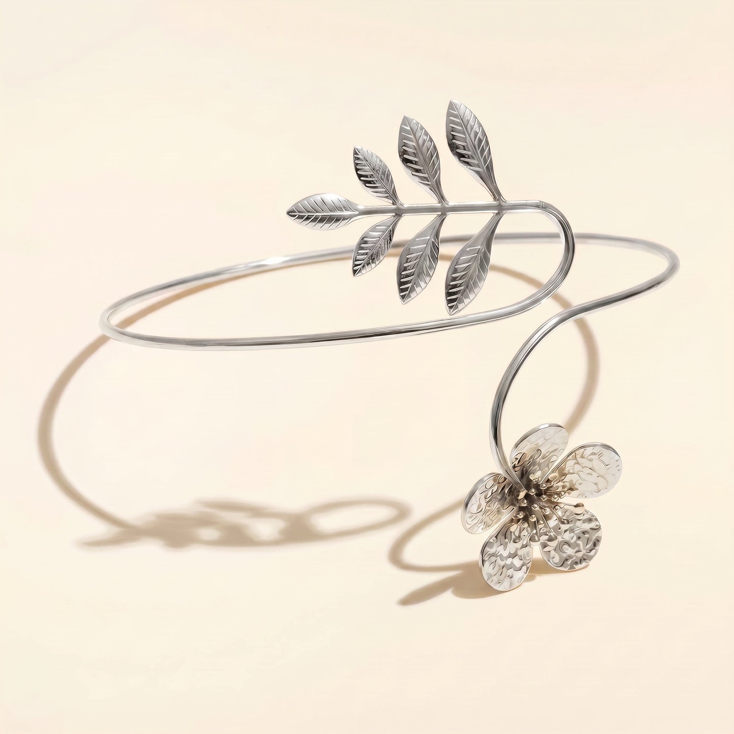 A silver open cuff bracelet featuring a leafy branch design and a dangling textured flower, displayed on a warm beige background with soft shadows.