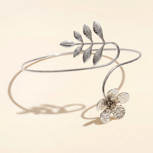 A silver open cuff bracelet featuring a leafy branch design and a dangling textured flower, displayed on a warm beige background with soft shadows.