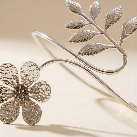 A silver-toned metal bangle bracelet featuring a textured five-petal flower and a leafy branch design, displayed on a warm beige background.