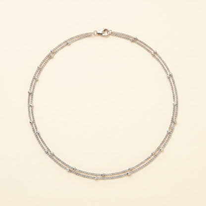 A delicate silver-toned double-strand necklace with small bead accents, displayed on a light cream background.