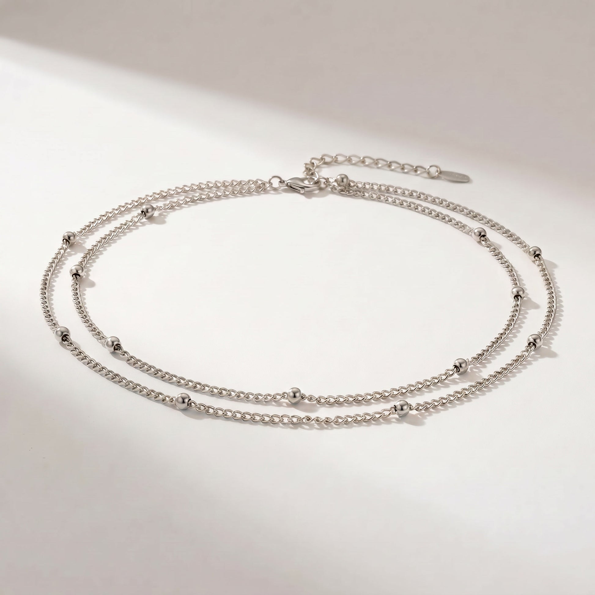 A silver double-layered chain anklet with small bead accents, displayed on a clean white surface with soft shadows.