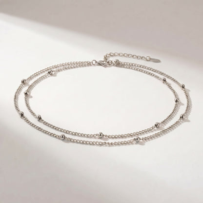 A silver double-layered chain anklet with small bead accents, displayed on a clean white surface with soft shadows.