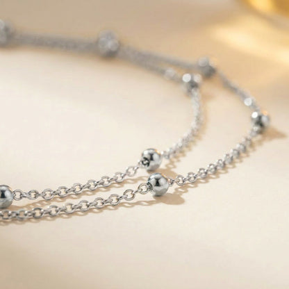 A close-up of a silver-toned double-strand chain necklace with polished spherical bead accents, resting on a soft beige surface.