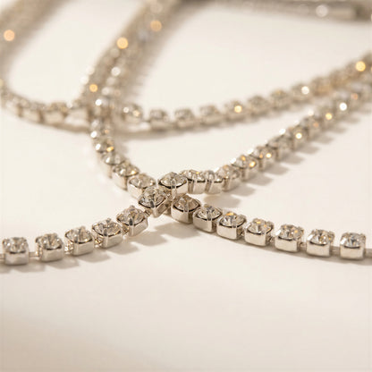 A close-up of a silver-toned tennis-style necklace with clear rhinestones arranged in a crisscross pattern on a light cream background.