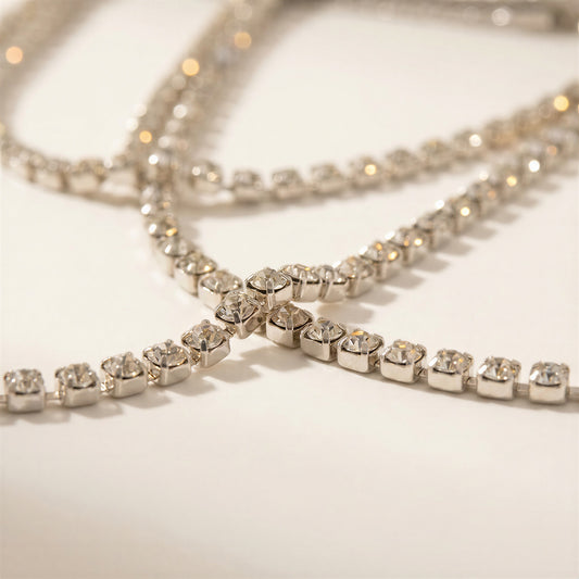 A close-up of a silver-toned tennis-style necklace with clear rhinestones arranged in a crisscross pattern on a light cream background.