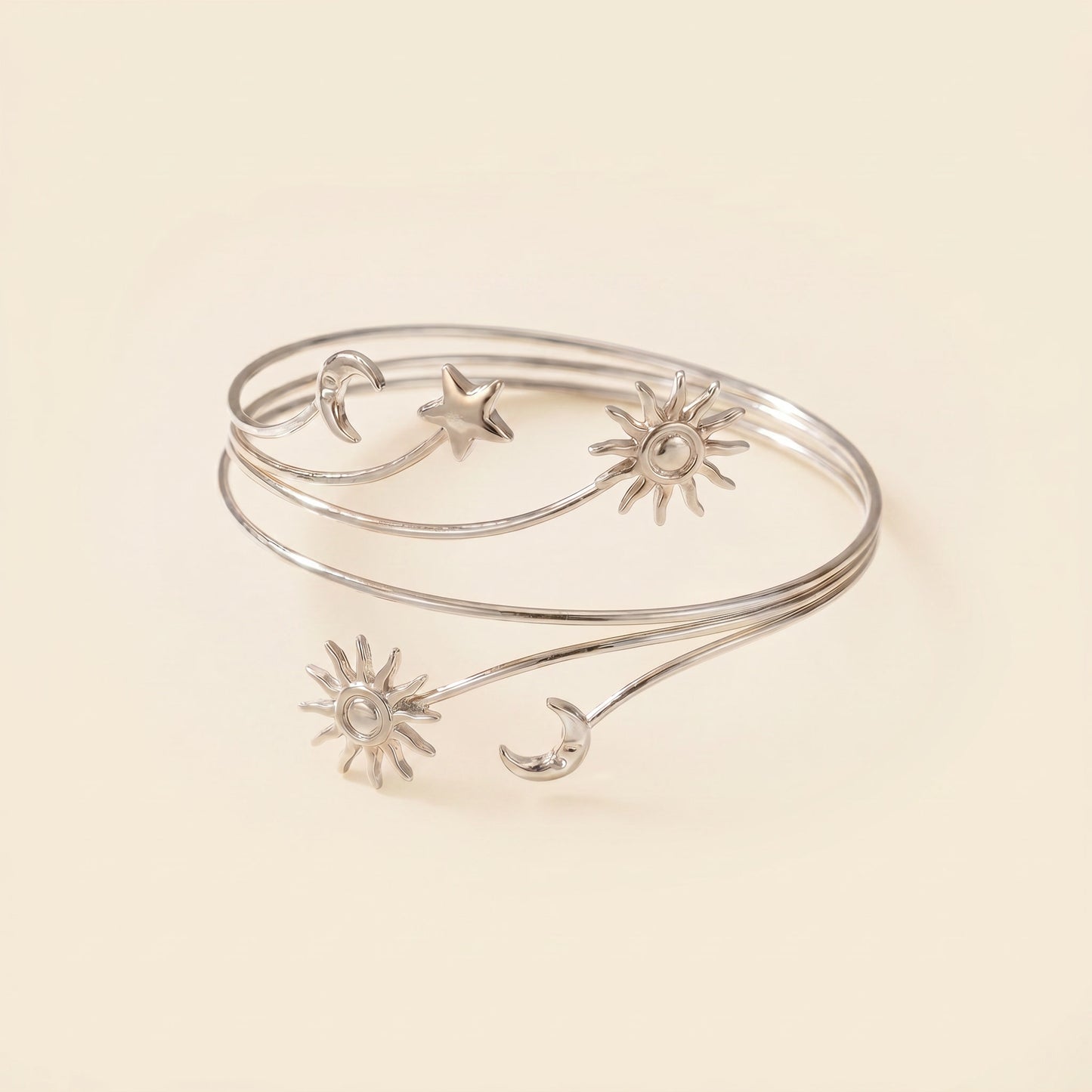A silver wrap bangle bracelet featuring delicate celestial motifs including suns, moons, and a star, displayed on a warm cream background.