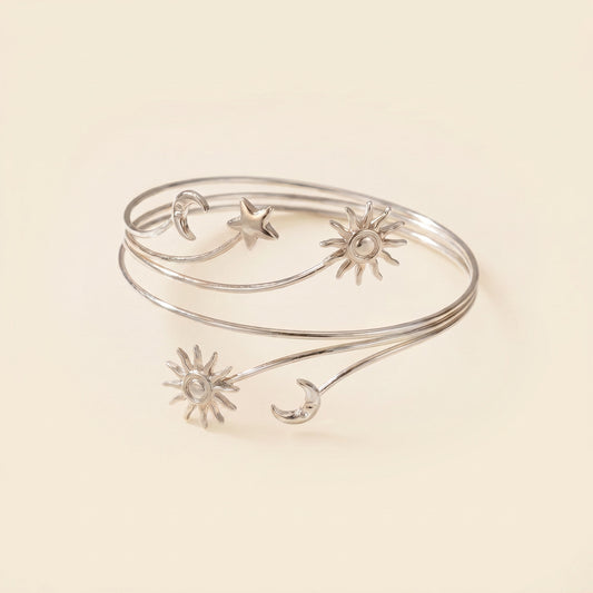 A silver wrap bangle bracelet featuring delicate celestial motifs including suns, moons, and a star, displayed on a warm cream background.