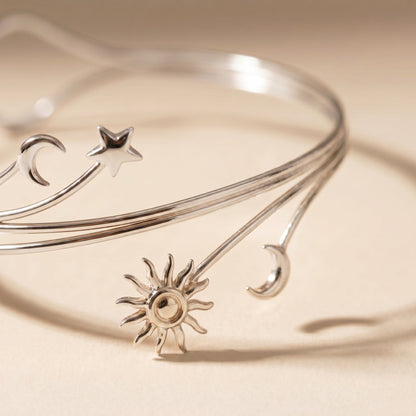 A polished silver bangle bracelet featuring celestial motifs, including a sun, crescent moons, and stars, displayed on a warm beige background.
