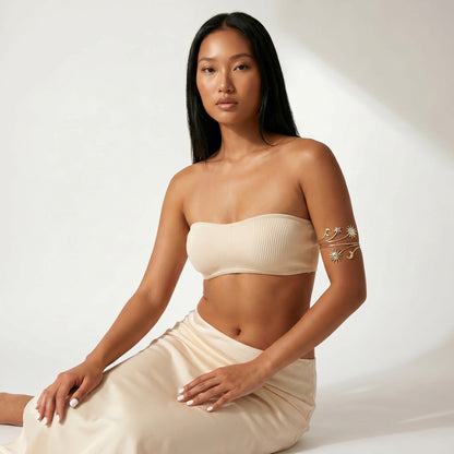 A woman wearing a beige ribbed strapless bandeau top and matching beige skirt, seated against a white studio background with soft diagonal shadows. She has a gold leaf-patterned armband accessory on her upper arm.