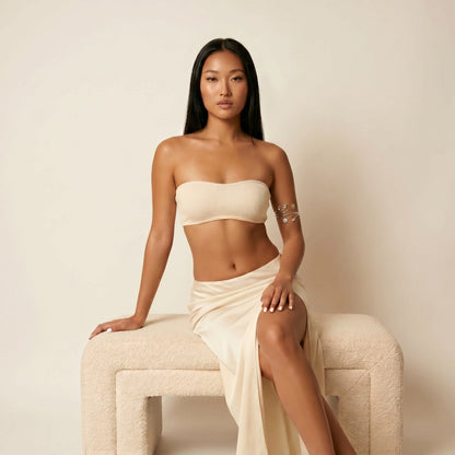 A woman with long black hair sits on a textured cream-colored bench against a solid cream background, wearing a matching beige ribbed bandeau top and draped maxi skirt.
