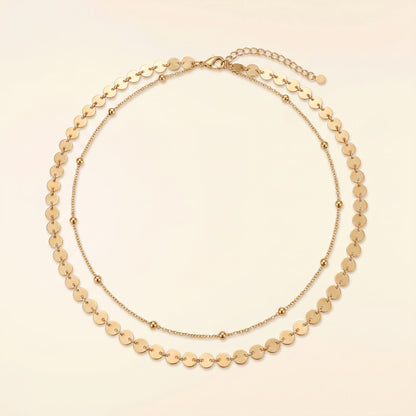 A gold-toned double-strand necklace featuring a layered design with flat circular disc links on the outer layer and smaller bead links on the inner layer, displayed on a light cream background.